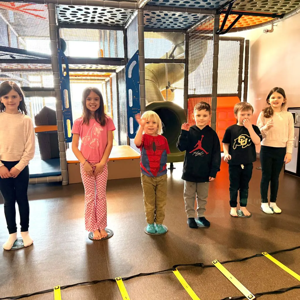 Kids standing in The Fitness Equation Kidz Club indoor play area with playground structure and agility ladder.
